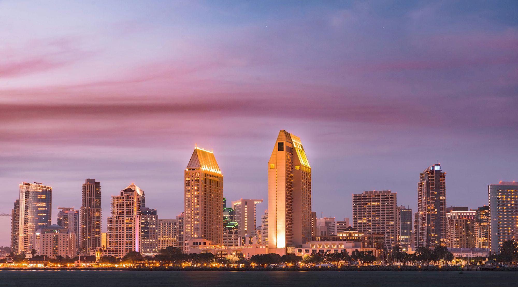 A city skyline at dusk with illuminated skyscrapers and a pink-purple sky, viewed across a body of water.