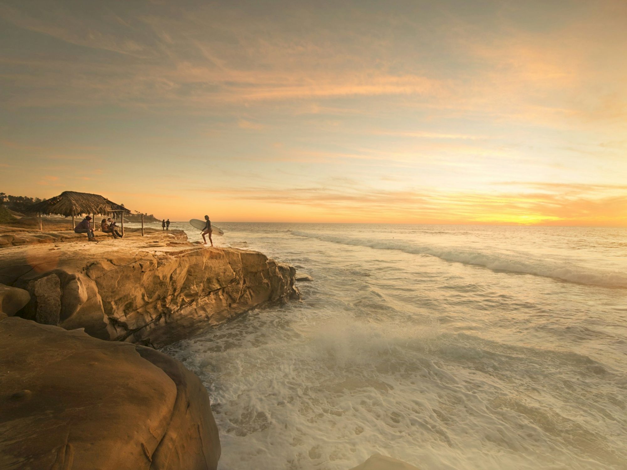 A surfer stands with a board on a rocky coastline at sunset, with onlookers nearby under a wooden shelter.