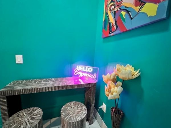 A cozy room with teal walls features a neon sign saying "Hello Gorgeous," vibrant artwork, rustic furniture, and decorative flowers.