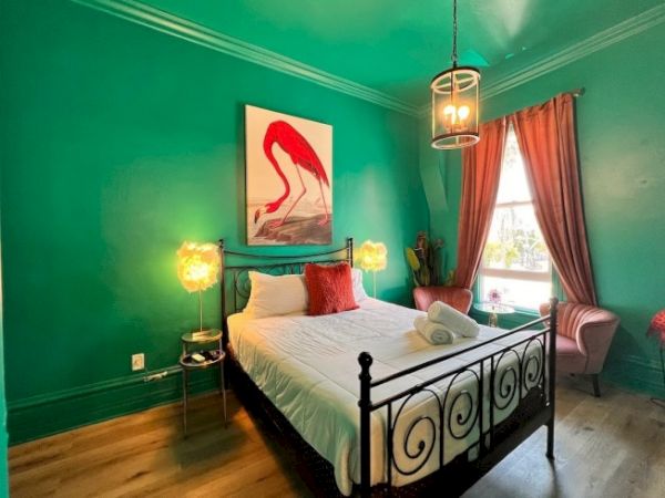 A vibrant bedroom with teal walls, metal bedframe, flamingo art, decorative lamps, and coral curtains, creating an eclectic cozy atmosphere.