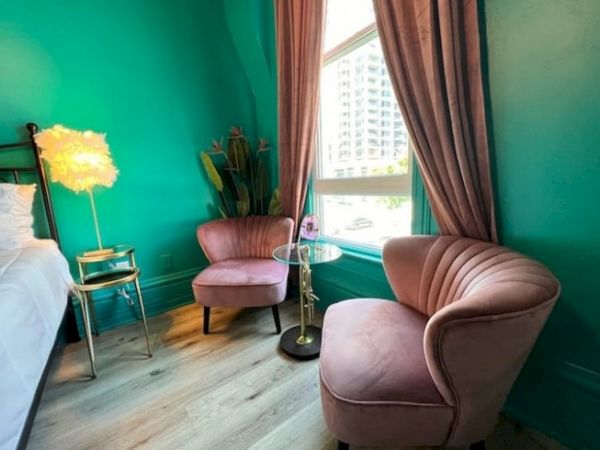 A cozy room features pink chairs, a fluffy lamp, a small table, vibrant teal walls, and a large window with a view of a building outside.