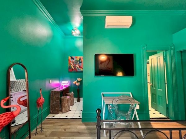 A vibrant room with teal walls, wall art, neon lights, a TV, a small table with a transparent chair, and flamingo decor.