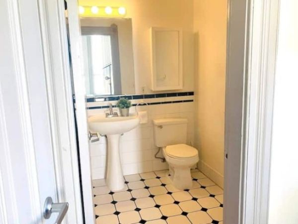 Small bathroom with white tiles, pedestal sink, toilet, and mirror. There’s a small plant on the sink and a bright overhead light.