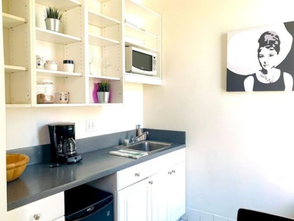 A small kitchen with open shelving, a coffee maker, microwave, and artwork on the wall. Minimalist design and decor.
