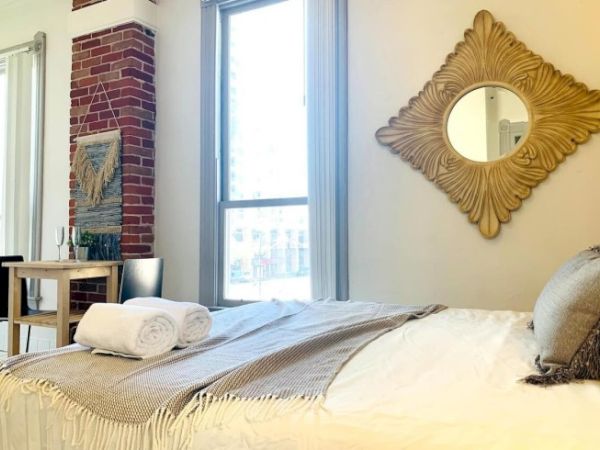 A cozy bedroom with a bed, two rolled towels, a decorative mirror, and a small table near an exposed brick wall.