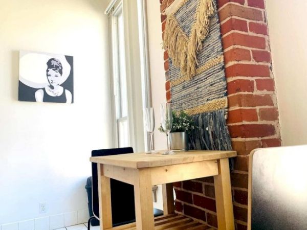 A cozy corner with a wooden table, chairs, brick wall, woven wall hanging, plant, and a monochrome portrait on the adjacent wall.