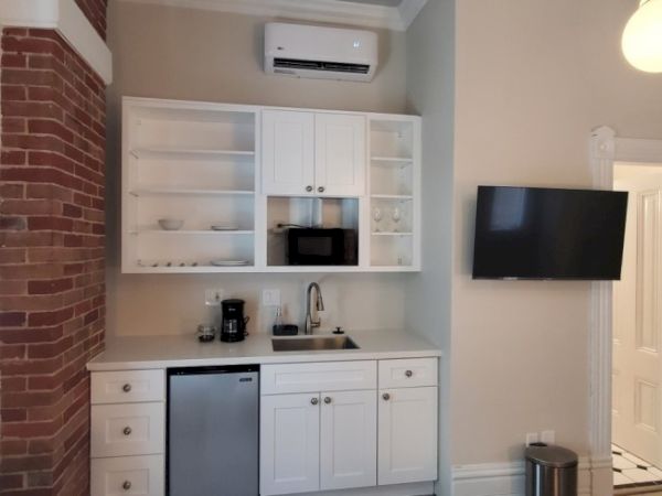 A compact kitchen with a sink, cabinets, a microwave, mini fridge, wall-mounted TV, and air conditioner, adjacent to a brick wall.