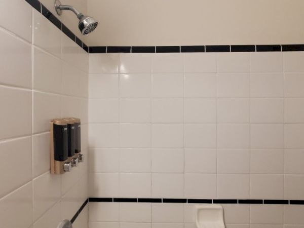 A tiled shower area with a showerhead and wall-mounted dispensers. The tiles are white with black border accents.
