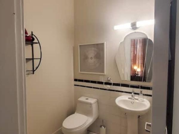 A small bathroom with a toilet, pedestal sink, mirror, light, wall art, and a small shelf on the left side.