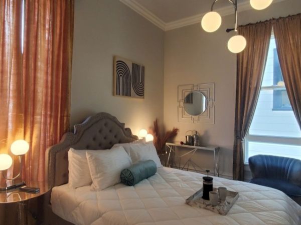 A cozy bedroom with a tufted bed, warm lighting, abstract art, and elegant curtains, featuring a chair and a small table by the window.