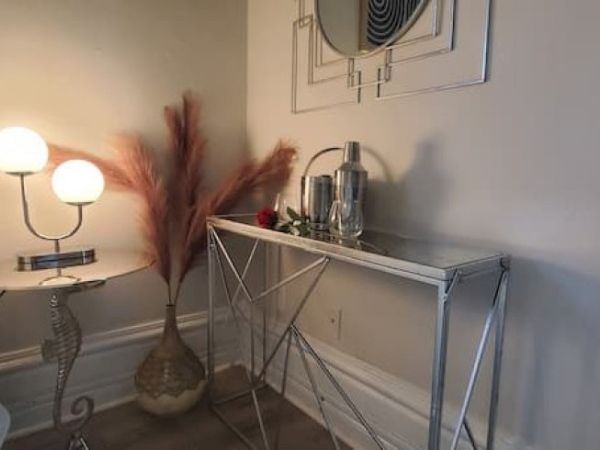 A modern side table with a metal frame holding a cocktail shaker and glasses; a round lamp and vase with pampas grass on the left.