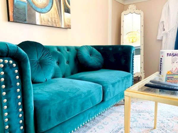 The image shows a teal tufted sofa with round cushions, a glass coffee table with a book and remote, plus a white standing mirror nearby.