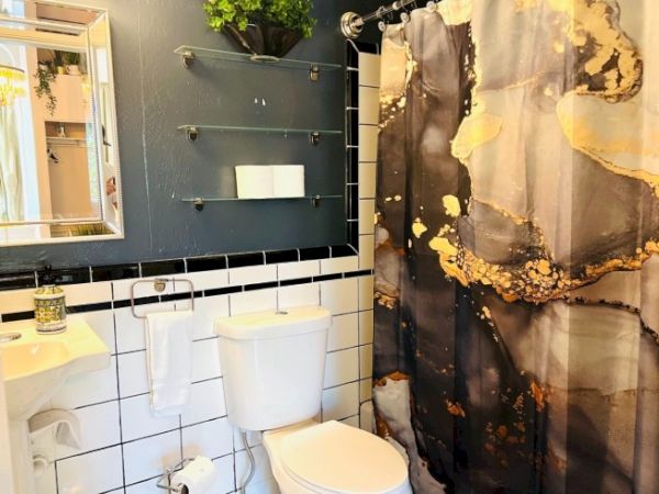A small bathroom with a modern design features a black and gold shower curtain, white tiled wall, toilet, sink, and shelving.