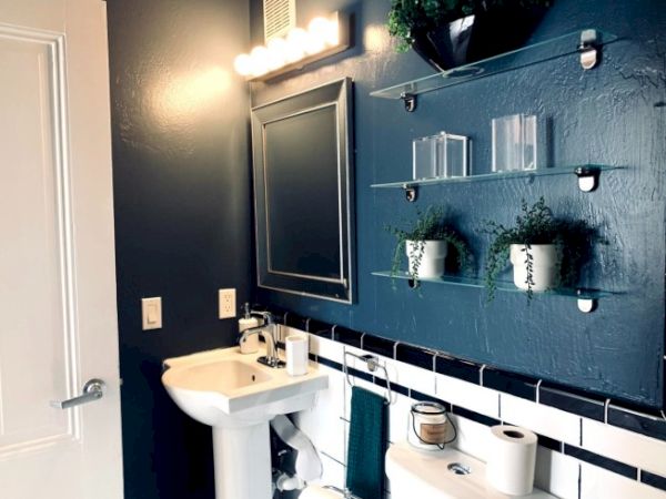 A bathroom with a sink, mirror, shelves with plants, toilet paper, and wall lights. The walls are dark, with a white and chrome theme.