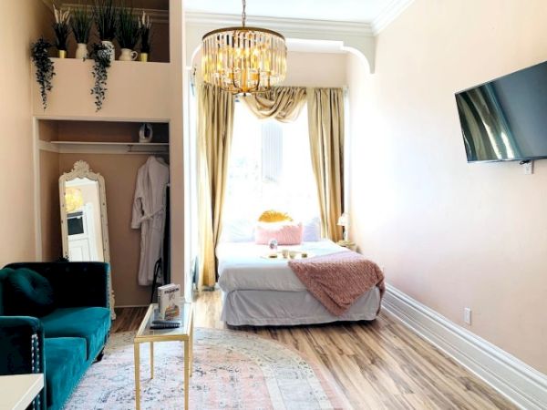 A cozy room with a bed, couch, wall-mounted TV, and plants, featuring warm colors and a chandelier for lighting.