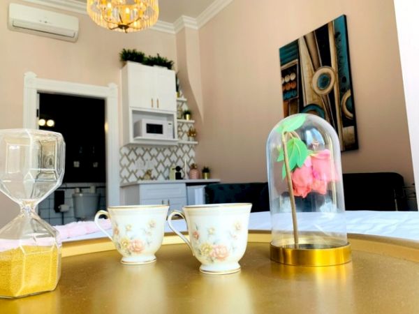 Decorative room with two floral teacups, an hourglass, and a rose in a glass dome on a golden table, modern kitchen in background.