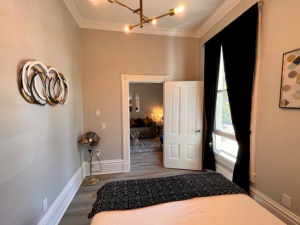 A room with a bed, wall art, a stylish fan, and a black curtain with a doorway leading to another room.