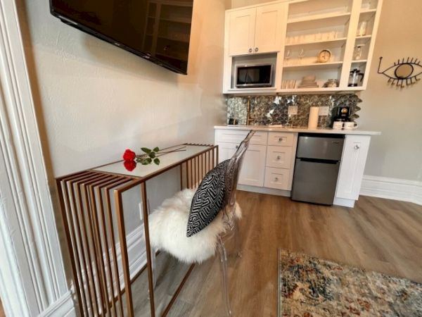 A kitchenette with white cabinets, a microwave, a small fridge, and a decorative wall. A table with a rose and a pillow on a chair is visible.