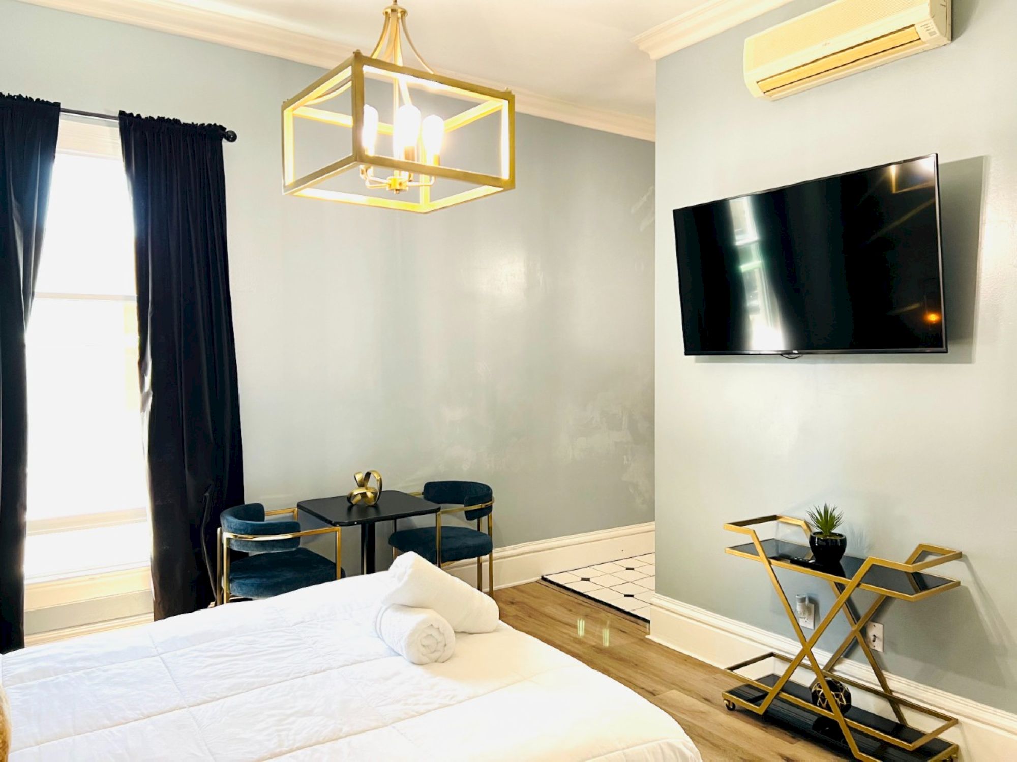 A modern bedroom features a bed, wall-mounted TV, chandelier, seating area, and a small table with a potted plant and towel on the bed.