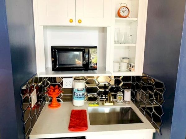 A compact kitchenette with a microwave, sink, hexagonal tile backsplash, orange decor, and a small shelf displaying a clock and mugs.