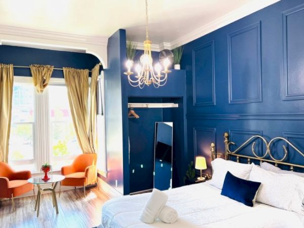 A stylish bedroom features a brass bed, deep blue walls, chandelier, seating area, and natural light from large windows with curtains.