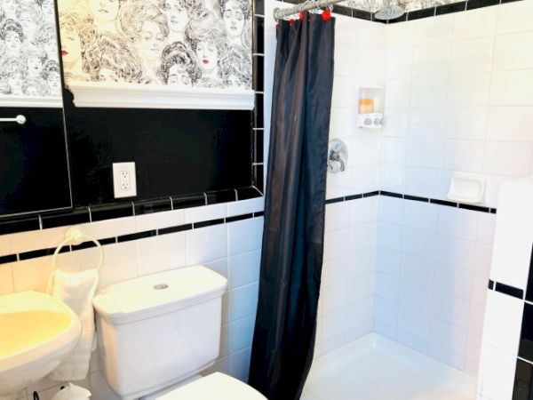 A bathroom with a sink, toilet, and shower. Black and white tiles, curtain, and wallpaper with illustrated faces complete the decor.