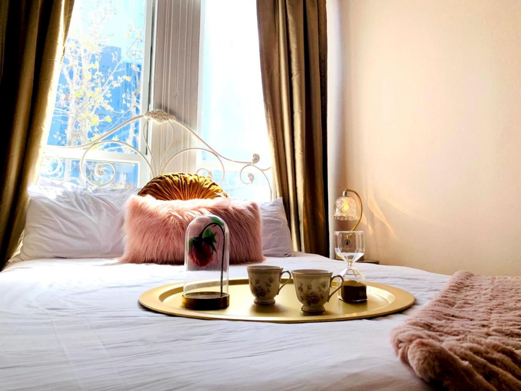 A cozy bedroom scene with a neatly made bed, decorative pillows, a tray with teacups, and a soft blanket draped at the end.