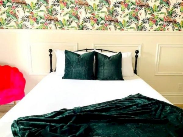 A bed with dark green pillows and blanket, white sheets, vibrant floral wallpaper, and a red chair in a bright room.