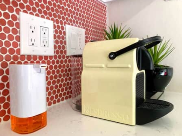 A coffee machine and soap dispenser on a counter with a red tiled backsplash, wall outlets, and small potted plants in the background.