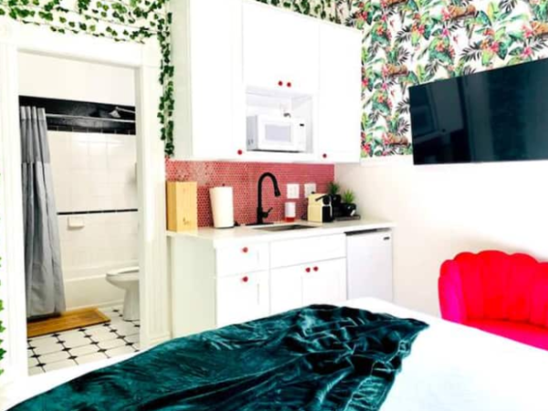 A bright room with tropical wallpaper, kitchenette, red chair, green blanket, and TV. Bathroom visible with a black-and-white tiled floor.