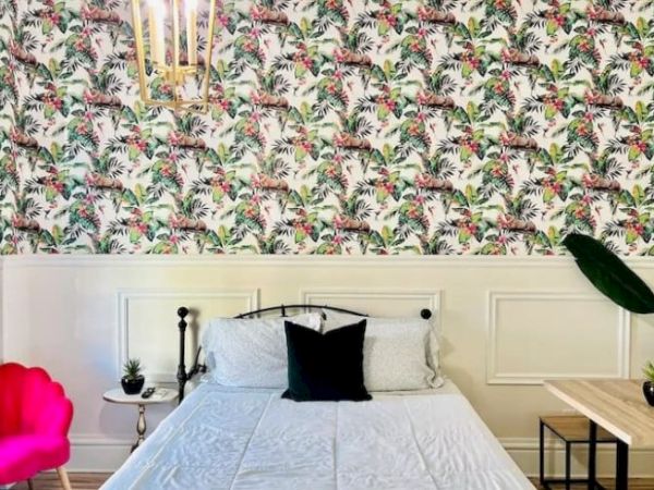 A bedroom with a colorful tropical wallpaper features a bed, black pillow, white bedding, side tables, plants, and a hanging light fixture.