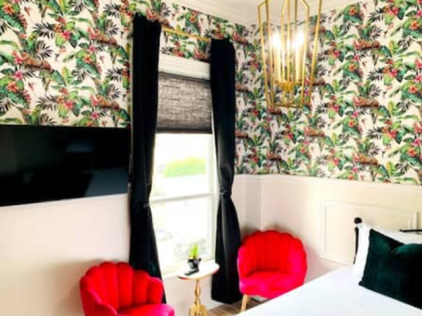 A room with tropical wallpaper, black curtains, red chairs, a white bed, a small table, a wall-mounted TV, and a modern light fixture.