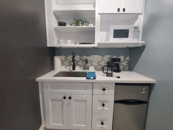 A small kitchenette featuring white cabinets, a sink, a microwave, a mini fridge, and a coffee maker on the countertop.