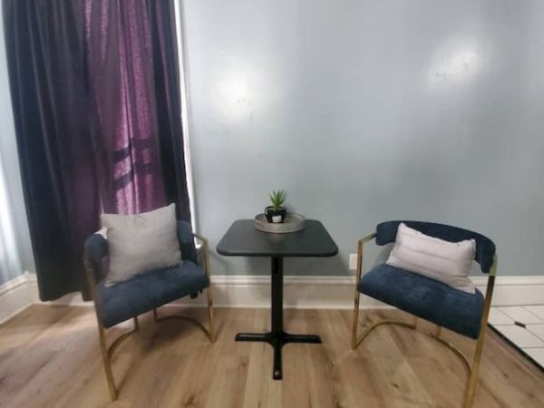 Two blue chairs flanking a small black table with a plant, in front of a purple-curtained window on a wooden floor.