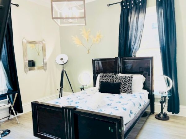 A bedroom with a bed, decorative pillows, standing lamps, a mirror, and dark curtains on windows.