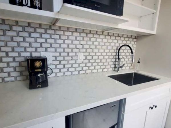 A kitchen countertop with a coffee maker, sink, and a modern backsplash, featuring white cabinetry and a microwave above.