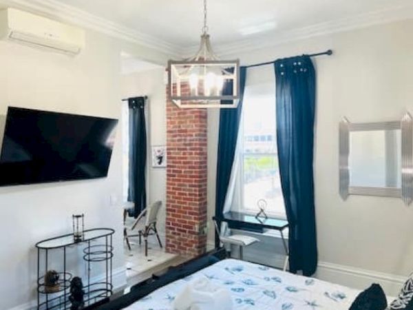 The image depicts a modern bedroom with a TV, a bed, a desk by the window, dark curtains, and a decorative brick column.