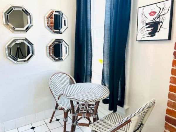 A small corner with three octagonal mirrors, wicker chairs, a round table, dark curtains, and wall art featuring a woman's face holding a cup.