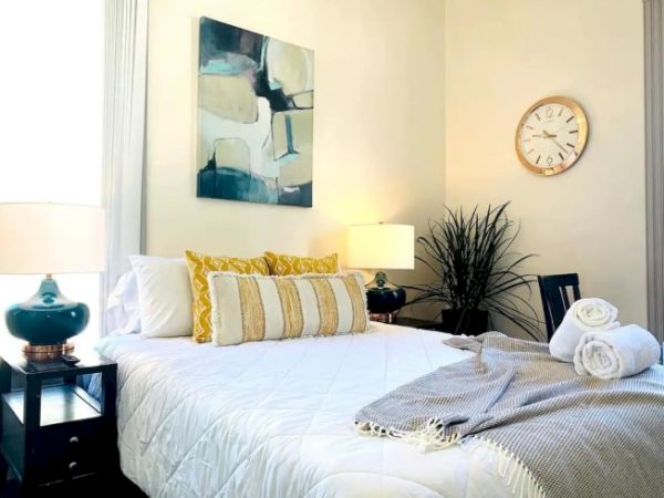 A cozy bedroom with a bed, abstract art, lamps, a clock, and neatly arranged towels.