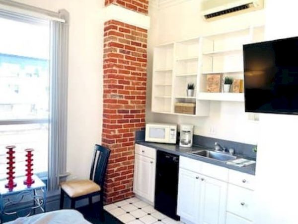 A small kitchen with white cabinets, a brick accent, and black countertop. Features include a microwave, coffee maker, sink, and wall-mounted TV.