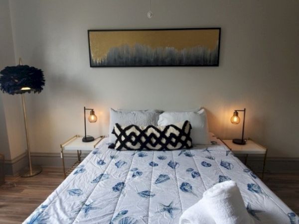 A cozy bedroom features a bed with blue-patterned bedding, artistic pillows, stylish lamps, and a modern artwork on the wall.