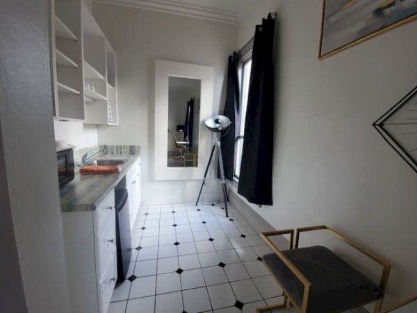 A narrow room with white cabinetry, checkered tile flooring, a large mirror, tripod lamp, dark curtains, and a cushioned chair.