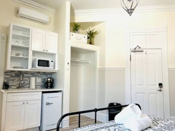 The image shows a room with a kitchenette, mini-fridge, microwave, bed, and neatly stacked towels on top, near a door and closet space.