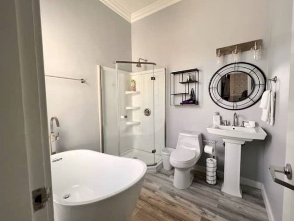 Modern bathroom with a freestanding tub, glass shower, toilet, pedestal sink, round mirror, shelves, light fixture, and wood-look floor.