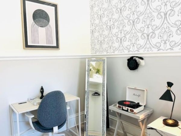 A room with a small desk, chair, floor mirror, record player, lamp, and abstract wall art. There's patterned wallpaper on one wall.