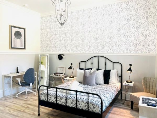 The image shows a stylish bedroom with a patterned wall, metal bed, desk, chair, and lamps, creating a cozy and modern atmosphere.