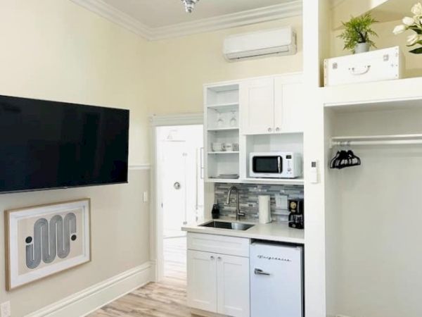 A compact kitchenette with a microwave, mini fridge, sink, coffee maker, and shelves. A large TV is mounted on the wall.