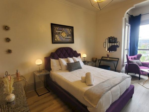 A stylish bedroom with a purple bed, modern art, chairs, and a large window with curtains, creating a cozy, elegant atmosphere.