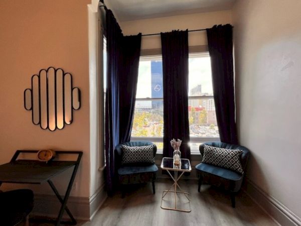 A cozy nook with two chairs, a small table, dark curtains, and a view of a cityscape through the window. A wall mirror decorates the space.