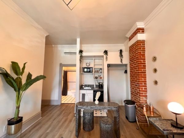 A cozy room with plants, a kitchenette, and a bathroom entrance. Features include a brick wall, small dining area, and wooden flooring.